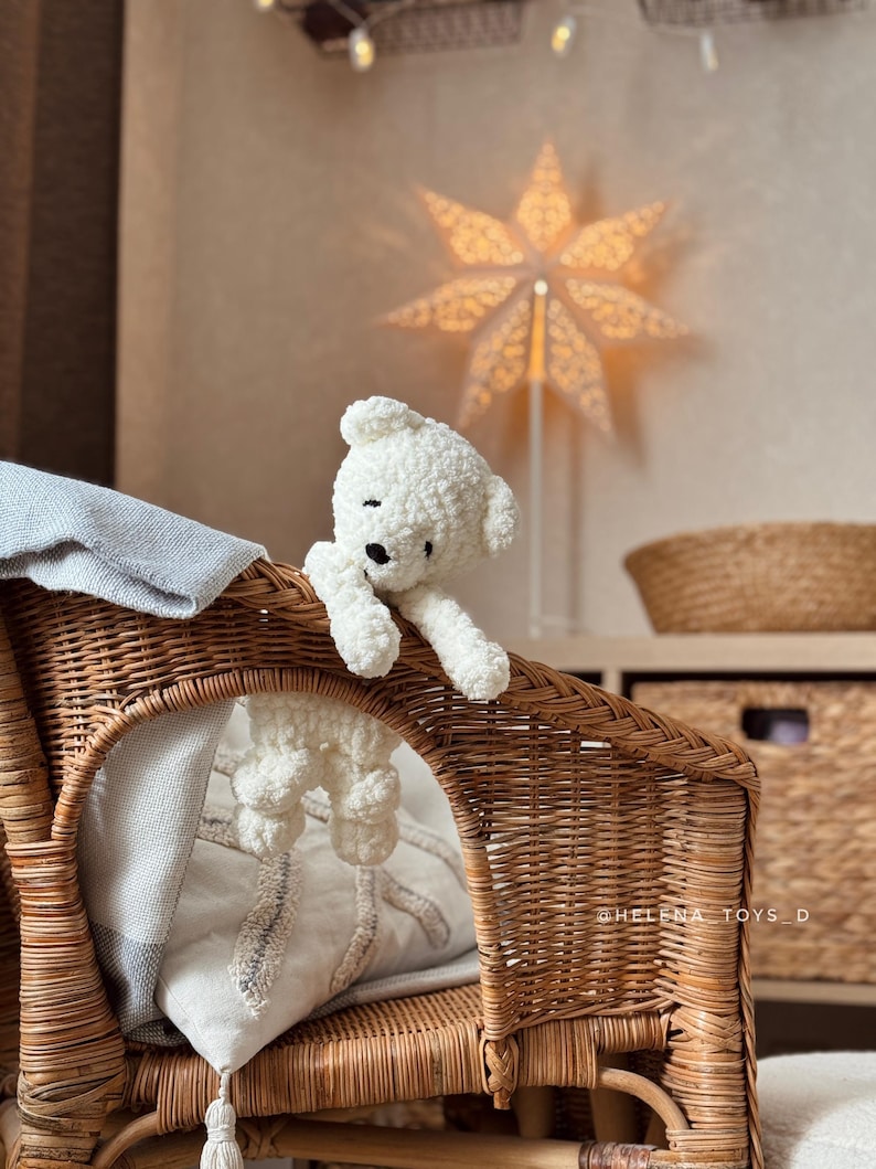 Pode incluir: Um urso de pel&uacute;cia branco feito &agrave; m&atilde;o com olhos pretos est&aacute; sentado em uma cadeira de vime. O urso est&aacute; pendurado na borda da cadeira. Uma lumin&aacute;ria em forma de estrela est&aacute; no fundo.