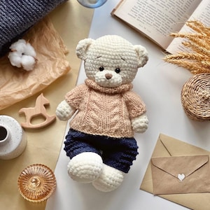 May include: A handmade, cream-colored crochet teddy bear wearing a light brown cable-knit sweater and navy blue shorts. The bear has a brown nose and is sitting on a neutral surface with other decorative items.