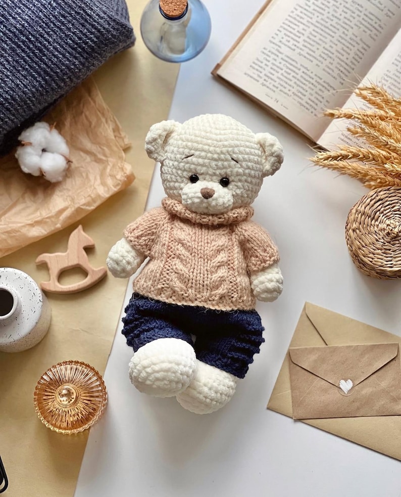May include: A handmade, cream-colored crochet teddy bear wearing a light brown cable-knit sweater and navy blue shorts. The bear has a brown nose and is sitting on a neutral surface with other decorative items.