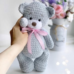 May include: A gray crocheted teddy bear with a pink ribbon around its neck. The bear has white fur around its eyes and a brown nose.