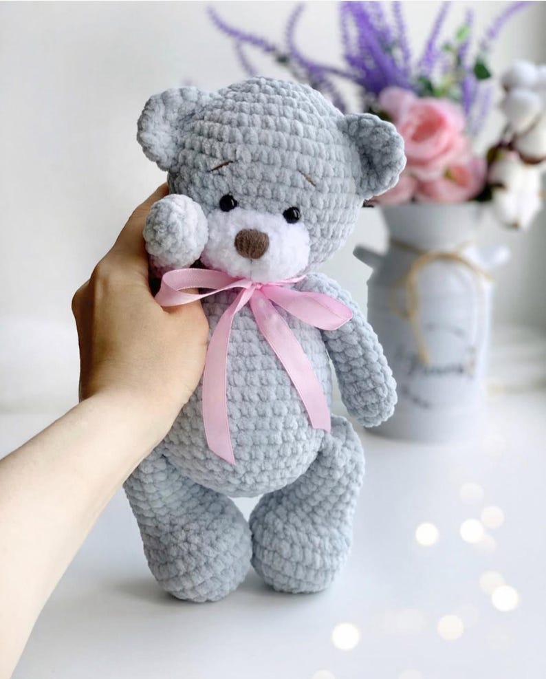 May include: A gray crocheted teddy bear with a pink ribbon around its neck. The bear has white fur around its eyes and a brown nose.