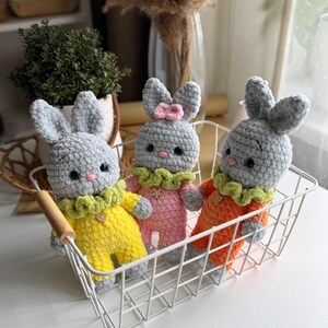 May include: Three crocheted gray bunny toys with different colored outfits. The bunnies are sitting in a white wire basket. The bunny on the left is wearing a yellow outfit with green trim, the bunny in the middle is wearing a pink outfit with green trim, and the bunny on the right is wearing an orange outfit with green trim.