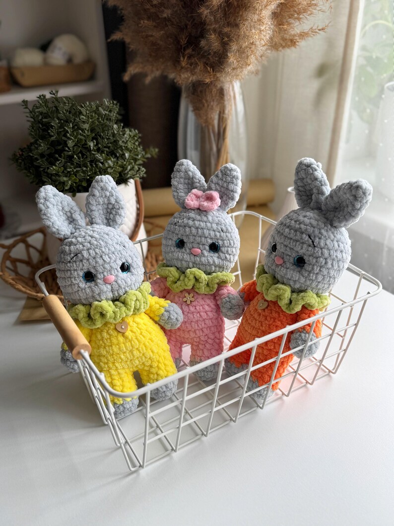 May include: Three crocheted gray bunny toys with different colored outfits. The bunnies are sitting in a white wire basket. The bunny on the left is wearing a yellow outfit with green trim, the bunny in the middle is wearing a pink outfit with green trim, and the bunny on the right is wearing an orange outfit with green trim.