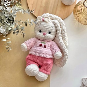 May include: A handmade crochet bunny doll with a white body, long floppy ears, and a pink bow. The bunny wears a pink sweater with cherry details and matching pink pants. The doll is a soft toy.