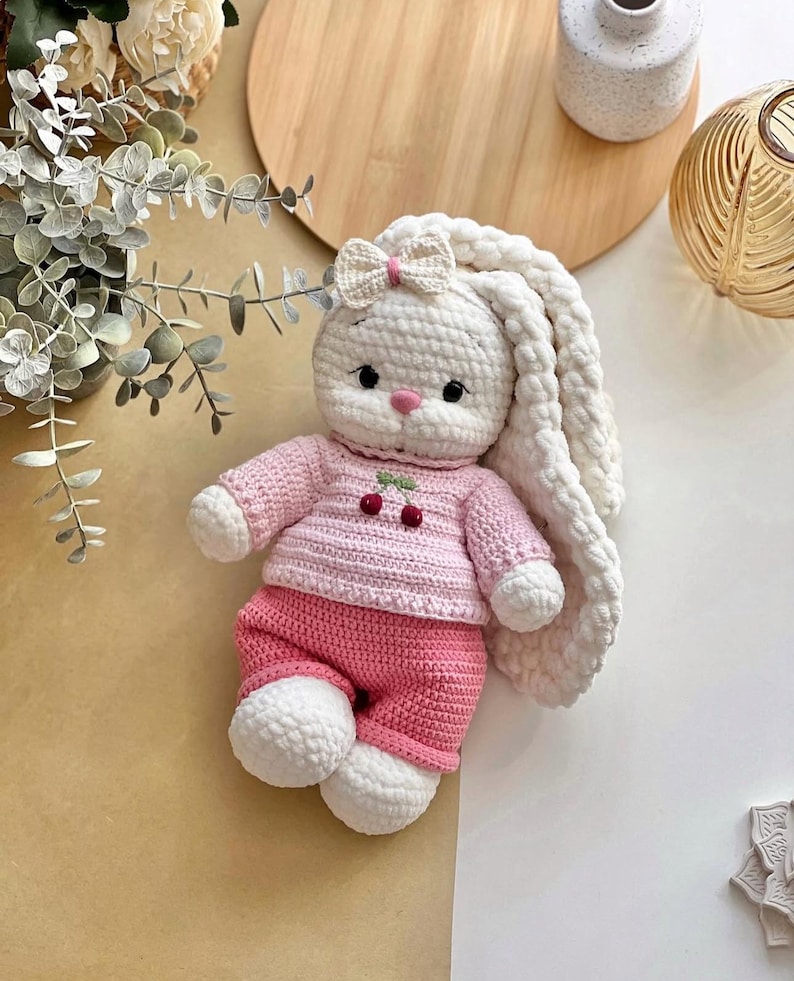 May include: A handmade crochet bunny doll with a white body, long floppy ears, and a pink bow. The bunny wears a pink sweater with cherry details and matching pink pants. The doll is a soft toy.