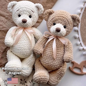 May include: Two handmade crochet teddy bears. One is off-white, the other is tan. Both have brown noses, black eyes, and pink ribbon bows. The bears are sitting side-by-side. Text in the image reads "Pattern crochet English" with an American flag.