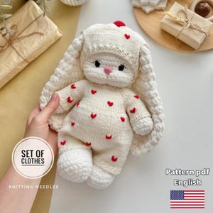 Puede incluir: Un muñeco de conejo tejido a mano, de color crema, con un gorro y un mono a juego, ambos adornados con pequeños corazones rojos. El conejo tiene orejas largas y caídas y una nariz rosa. El texto dice "SET OF CLOTHES" y "Pattern pdf English".