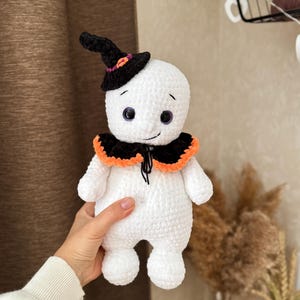 May include: A handmade white crochet ghost doll wearing a black witch's hat with orange and purple details. The ghost has a black and orange collar and purple eyes. The doll is being held.