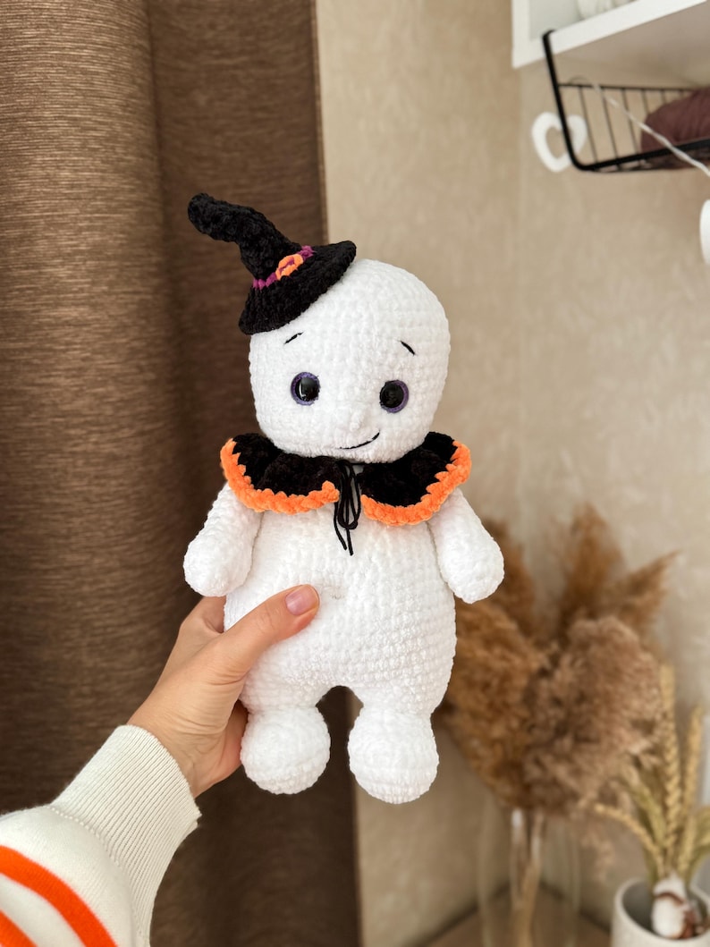 May include: A handmade white crochet ghost doll wearing a black witch's hat with orange and purple details. The ghost has a black and orange collar and purple eyes. The doll is being held.