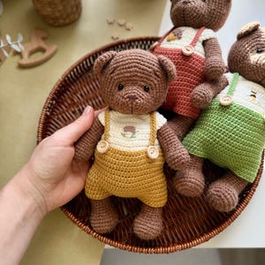 May include: Crocheted teddy bears in a brown wicker basket. The bears are brown with different colored overalls: yellow, red, and green. Each bear has a white shirt with a small embroidered design. The bears are handmade and have button details.