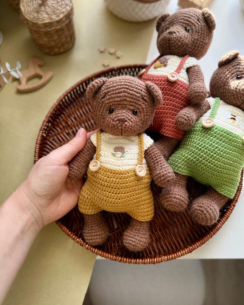 May include: Crocheted teddy bears in a brown wicker basket. The bears are brown with different colored overalls: yellow, red, and green. Each bear has a white shirt with a small embroidered design. The bears are handmade and have button details.