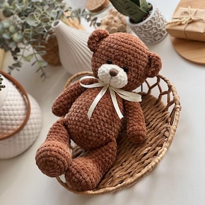 May include: A brown crocheted teddy bear with a white face and a white satin ribbon around its neck. The bear is sitting in a woven basket.