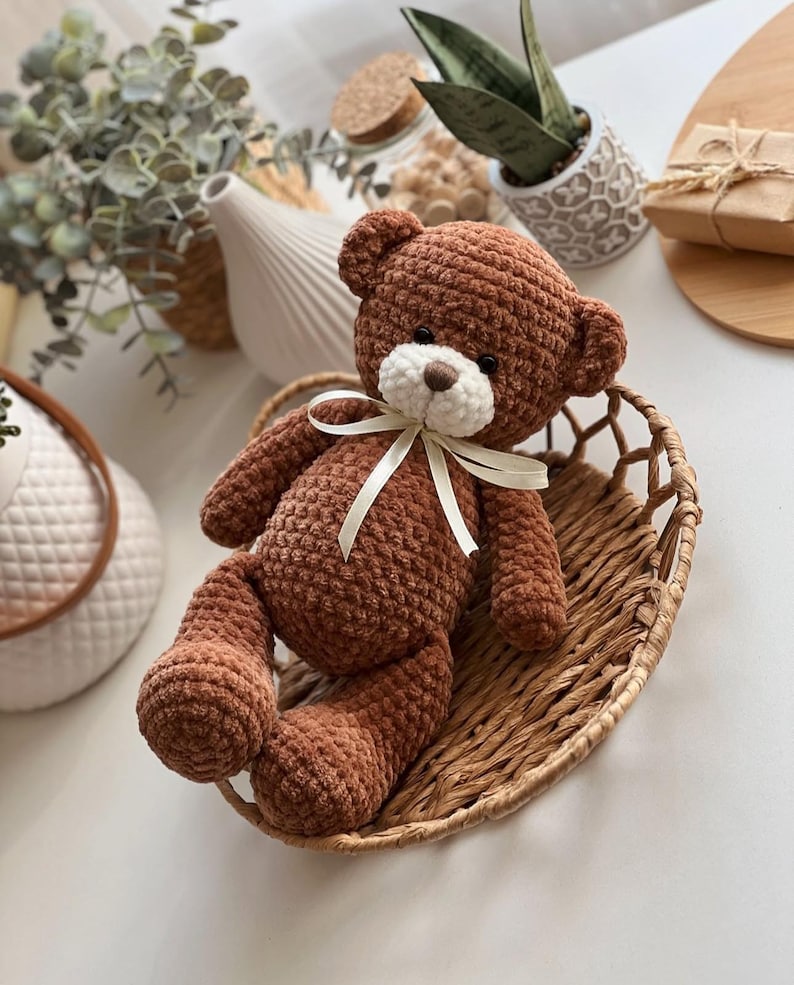 May include: A brown crocheted teddy bear with a white face and a white satin ribbon around its neck. The bear is sitting in a woven basket.