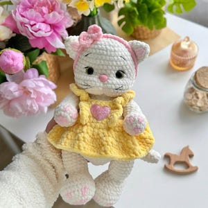 Pode incluir: Boneca de gato de croch&ecirc; branco feita &agrave; m&atilde;o, vestindo um vestido amarelo com um cora&ccedil;&atilde;o rosa e uma faixa de la&ccedil;o rosa. A boneca tem detalhes em rosa nas patas e orelhas. &Eacute; um brinquedo macio.