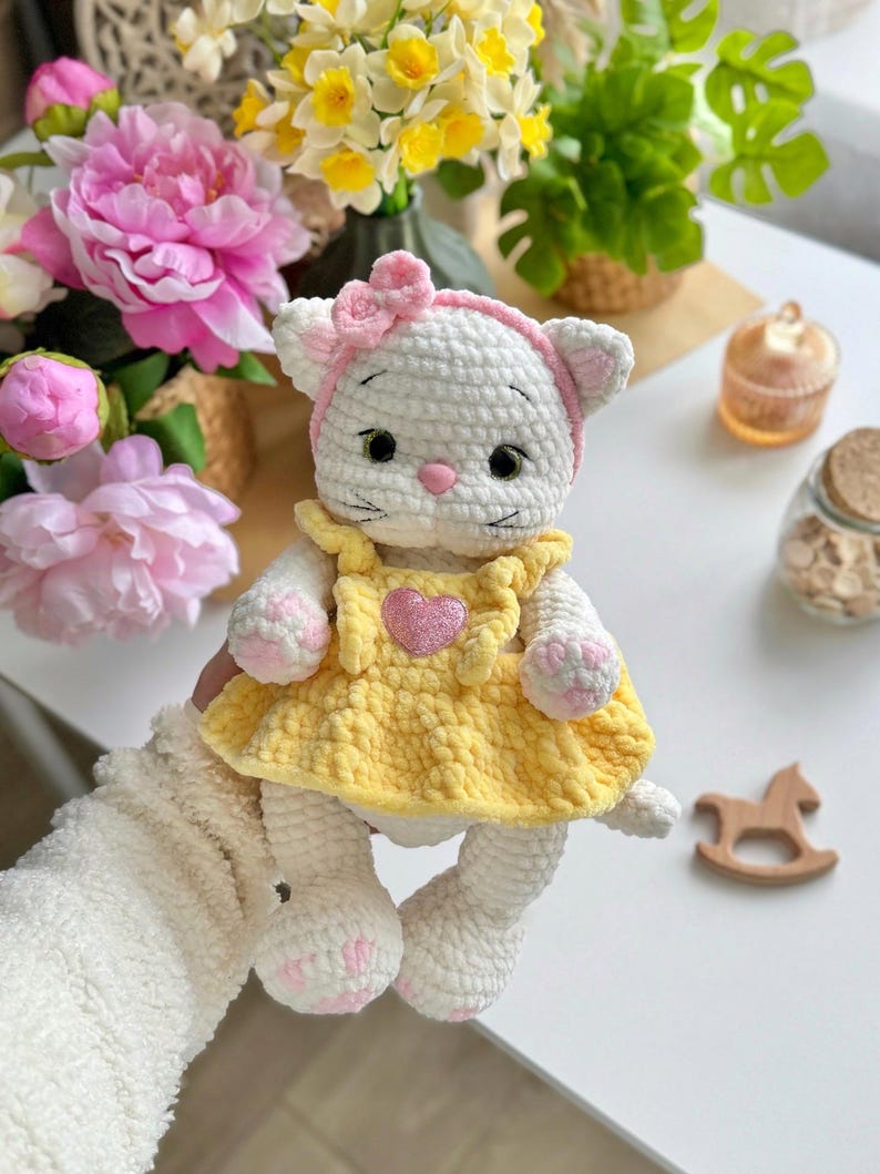Pode incluir: Boneca de gato de croch&ecirc; branco feita &agrave; m&atilde;o, vestindo um vestido amarelo com um cora&ccedil;&atilde;o rosa e uma faixa de la&ccedil;o rosa. A boneca tem detalhes em rosa nas patas e orelhas. &Eacute; um brinquedo macio.