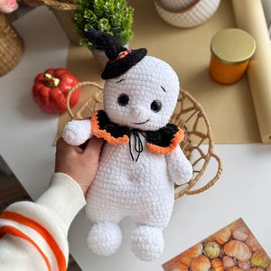 May include: A handmade white crochet ghost doll wearing a black witch hat with an orange trim and a black and orange collar. The ghost has black button eyes and a stitched smile. The doll is holding a basket.