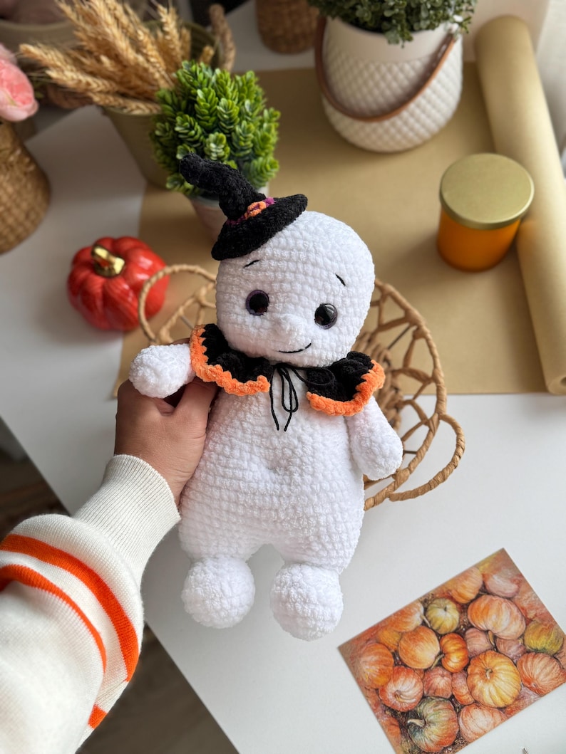 May include: A handmade white crochet ghost doll wearing a black witch hat with an orange trim and a black and orange collar. The ghost has black button eyes and a stitched smile. The doll is holding a basket.