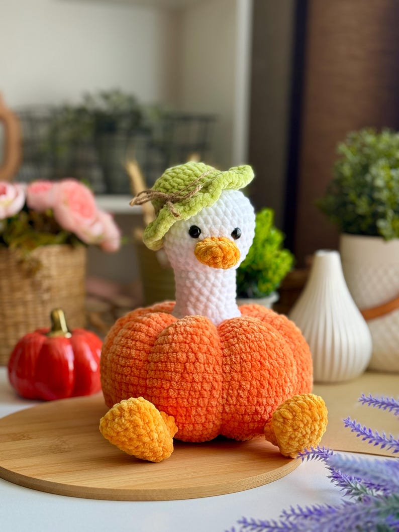 May include: A handmade crochet goose with a white head, orange beak, and a green hat. The goose is attached to an orange pumpkin body. Two small orange crochet pumpkins are on a wooden surface. The background includes plants and decorative items.