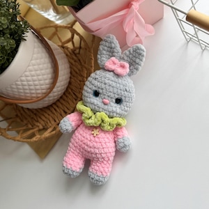 May include: A gray crocheted bunny with pink and green accents. The bunny is wearing a pink and gray outfit with a green ruffle collar and a pink bow on its head.