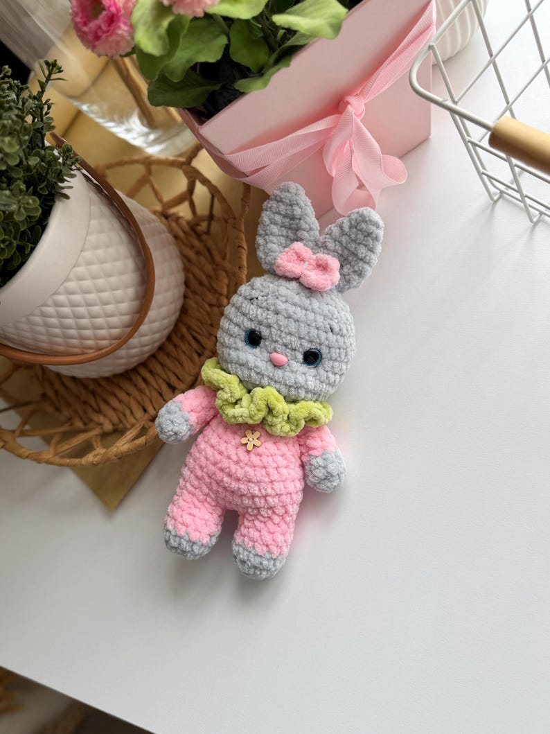 May include: A gray crocheted bunny with pink and green accents. The bunny is wearing a pink and gray outfit with a green ruffle collar and a pink bow on its head.