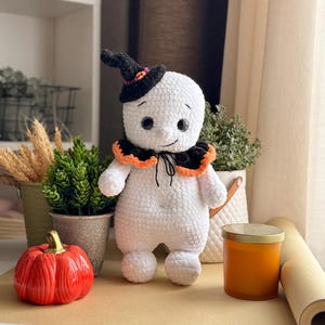 May include: A handmade white crochet ghost doll wearing a black witch's hat with orange and pink details. The ghost has a black collar with orange trim and black button eyes. An orange pumpkin and other decorations are in the background.