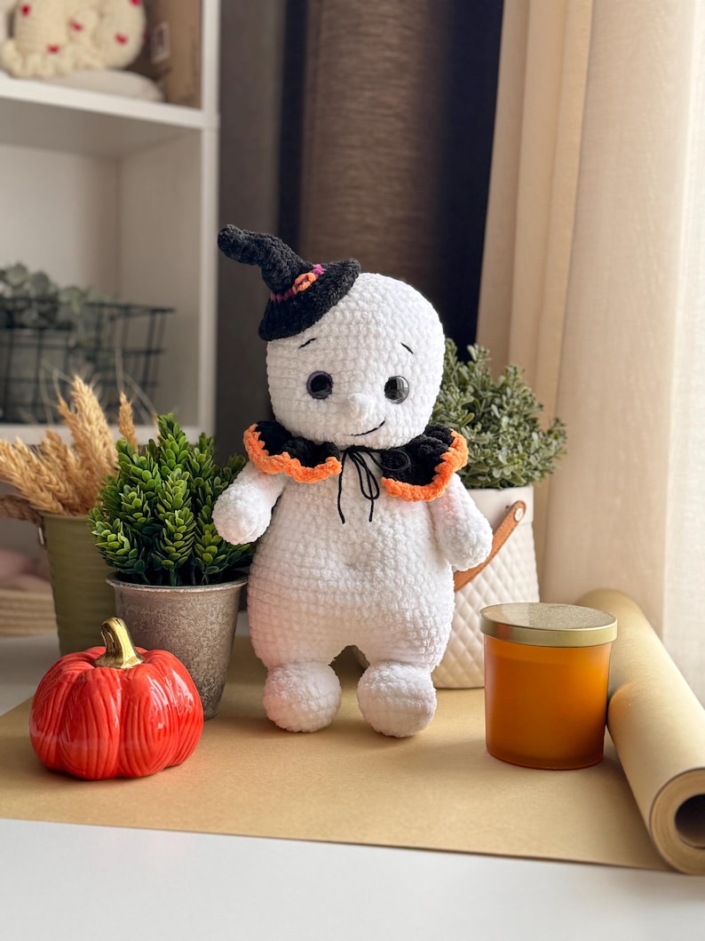 May include: A handmade white crochet ghost doll wearing a black witch's hat with orange and pink details. The ghost has a black collar with orange trim and black button eyes. An orange pumpkin and other decorations are in the background.