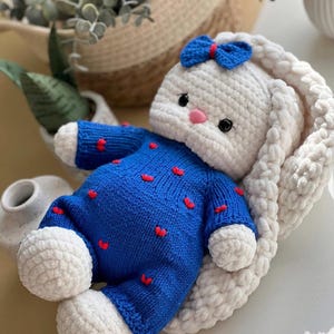 Puede incluir: Una mu&ntilde;eca de conejo hecha a mano con ganchillo. El conejo es blanco con un lazo azul y una nariz rosa. Viste un traje azul con detalles de corazones rojos. El conejo est&aacute; tumbado sobre una superficie suave y blanca.