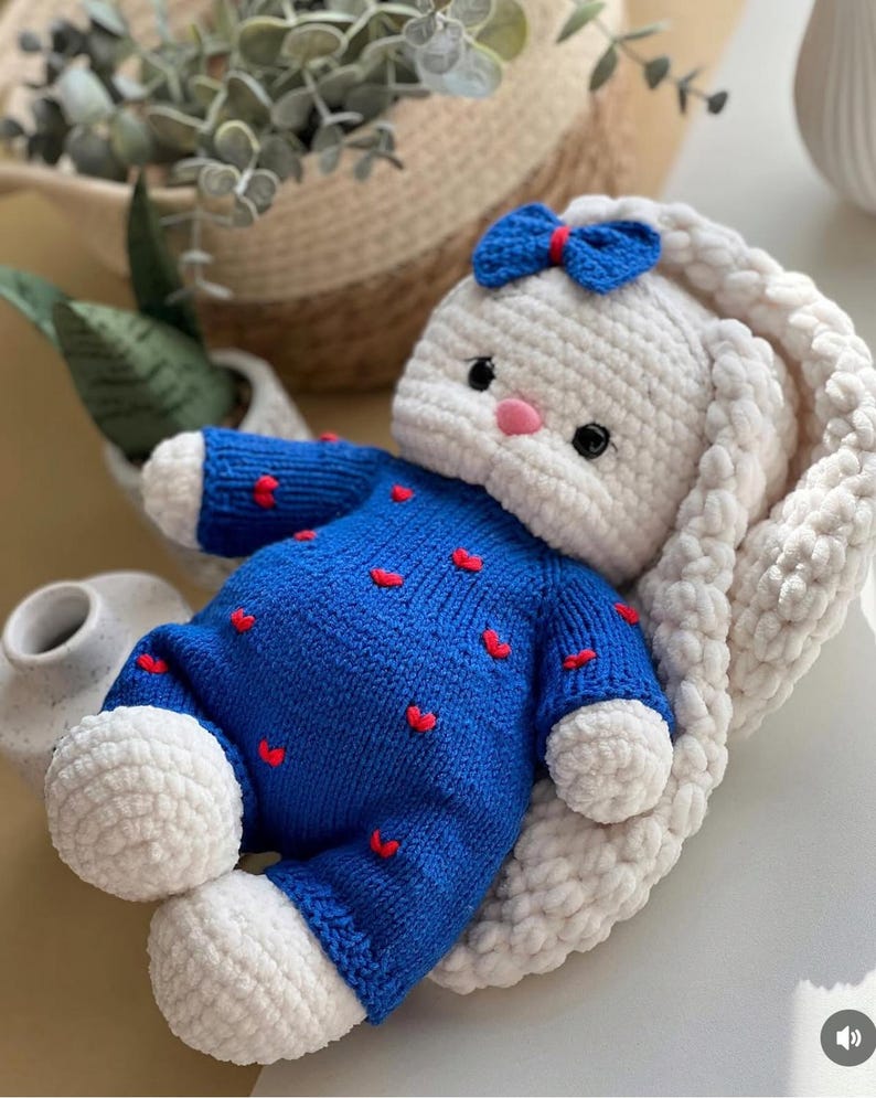 Puede incluir: Una mu&ntilde;eca de conejo hecha a mano con ganchillo. El conejo es blanco con un lazo azul y una nariz rosa. Viste un traje azul con detalles de corazones rojos. El conejo est&aacute; tumbado sobre una superficie suave y blanca.