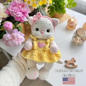 Może przedstawiać: Ręcznie robiona biała szydełkowa lalka kota w żółtej sukience z różowym sercem. Kot ma różową kokardę i opaskę na głowę. Lalka trzyma różowy i biały kwiat. Obraz zawiera tekst "Pattern crochet English".