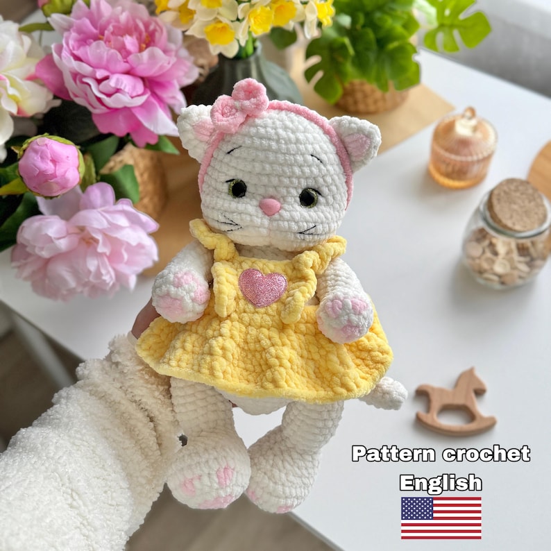 Pode incluir: Uma boneca de gato de croch&ecirc; branco feita &agrave; m&atilde;o usando um vestido amarelo com um cora&ccedil;&atilde;o rosa. O gato tem um la&ccedil;o e uma faixa de cabelo rosa. A boneca est&aacute; segurando uma flor rosa e branca. A imagem inclui o texto "Pattern crochet English".