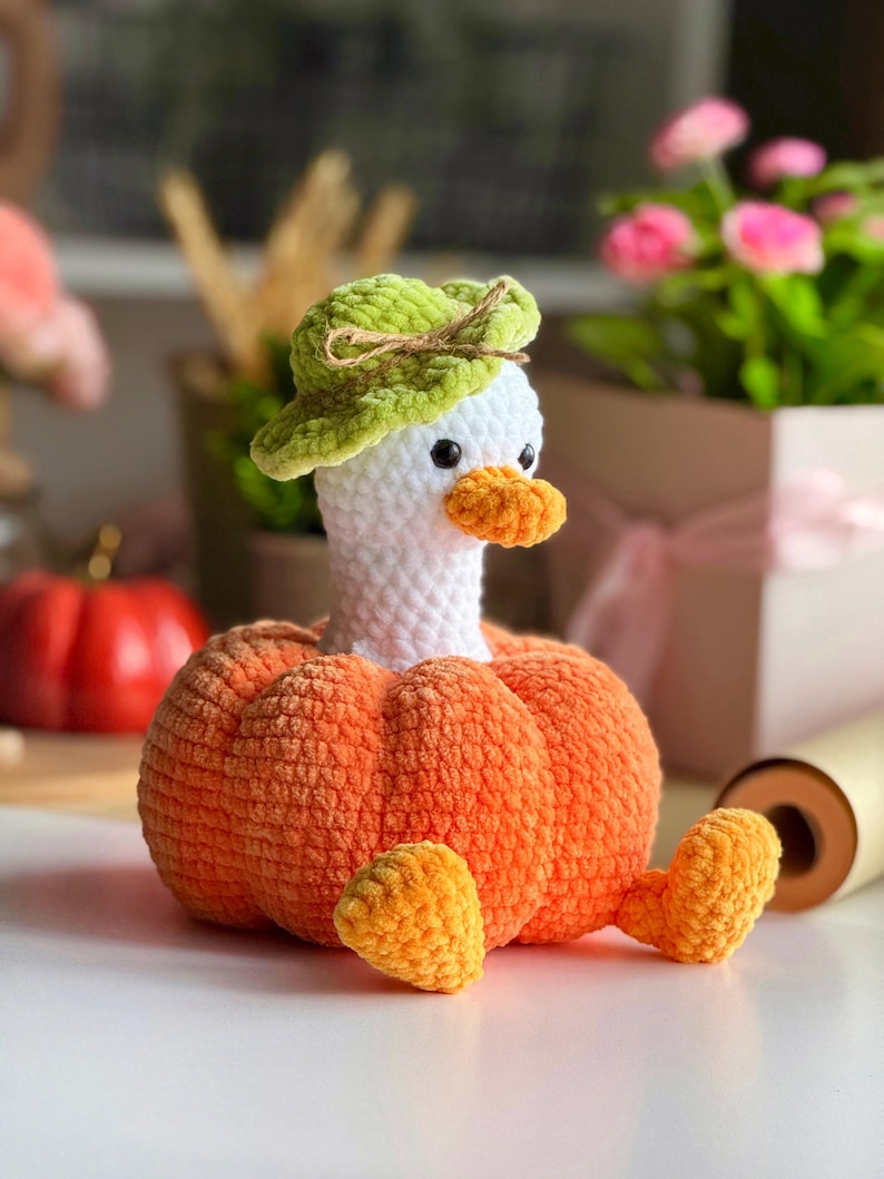 May include: A handmade crochet duck wearing a green hat, with an orange beak and feet, and a pumpkin-shaped body. The duck is white, and the pumpkin is a vibrant orange. The craft is likely for fall or Halloween decor.