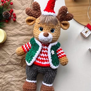 May include: A handmade crochet reindeer doll wearing a red and white striped sweater, green vest, and gray pants. The reindeer has brown antlers, a red Santa hat, and brown boots. A festive holiday decoration.