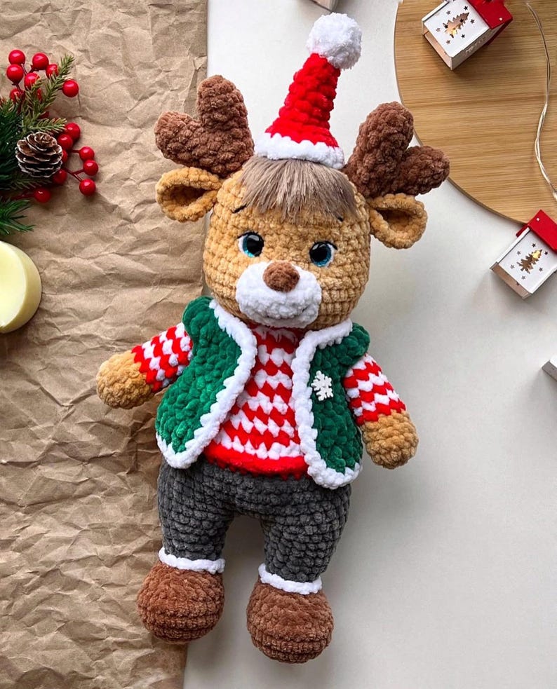 May include: A handmade crochet reindeer doll wearing a red and white striped sweater, green vest, and gray pants. The reindeer has brown antlers, a red Santa hat, and brown boots. A festive holiday decoration.