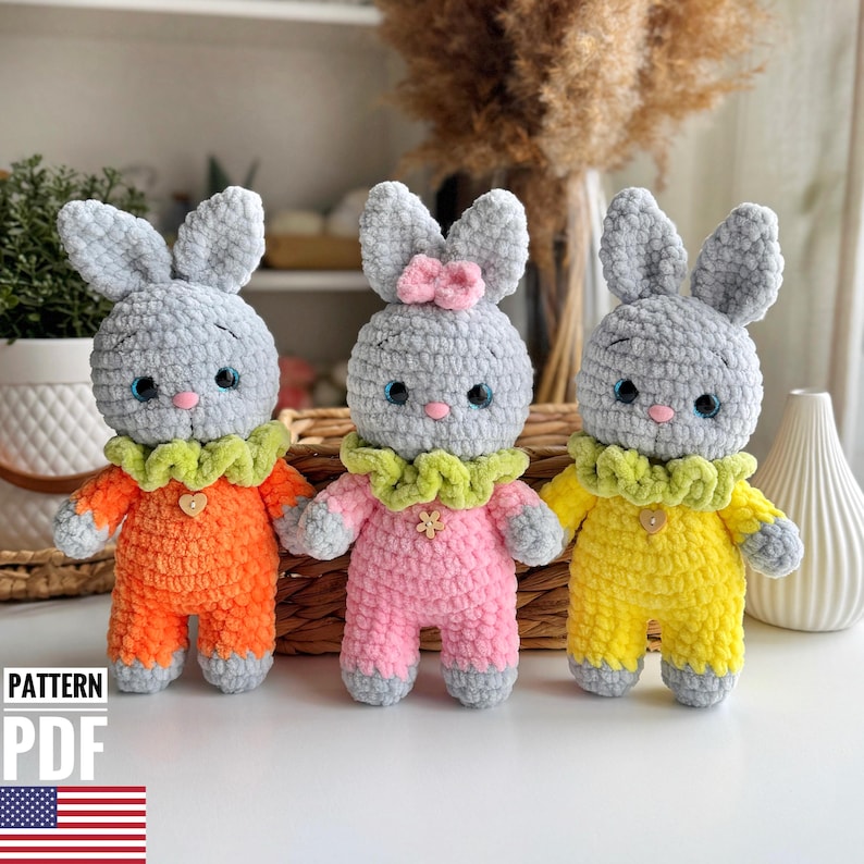 May include: Three crocheted gray bunny toys wearing different colored outfits. The bunny on the left is wearing orange, the middle bunny is wearing pink, and the bunny on the right is wearing yellow.