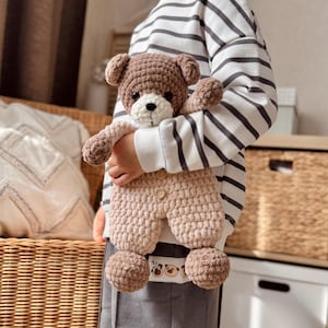 May include: A child holding a brown and beige crocheted teddy bear wearing a brown and beige crocheted outfit. The teddy bear has a white face with black eyes and a black nose. The child is wearing a striped white and grey sweater and grey pants.
