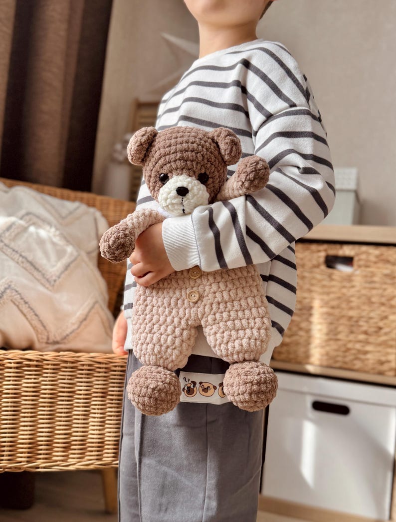 May include: A child holding a brown and beige crocheted teddy bear wearing a brown and beige crocheted outfit. The teddy bear has a white face with black eyes and a black nose. The child is wearing a striped white and grey sweater and grey pants.