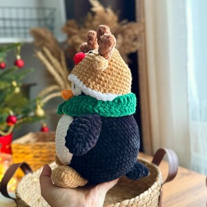 May include: A handmade crochet penguin wearing a reindeer hat and a green scarf. The penguin has a black body, white belly, and tan feet. The reindeer hat is tan with brown antlers and a red nose. The penguin is sitting in a woven basket.