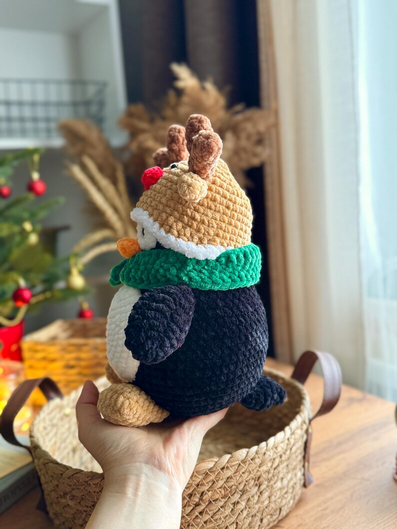 May include: A handmade crochet penguin wearing a reindeer hat and a green scarf. The penguin has a black body, white belly, and tan feet. The reindeer hat is tan with brown antlers and a red nose. The penguin is sitting in a woven basket.