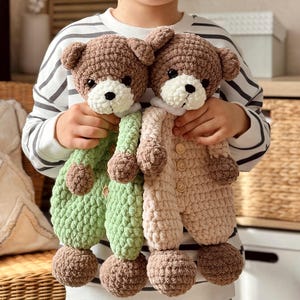 May include: Two crocheted teddy bears wearing matching outfits. The bear on the left is wearing a green outfit and the bear on the right is wearing a beige outfit. Both bears have brown fur and white faces.