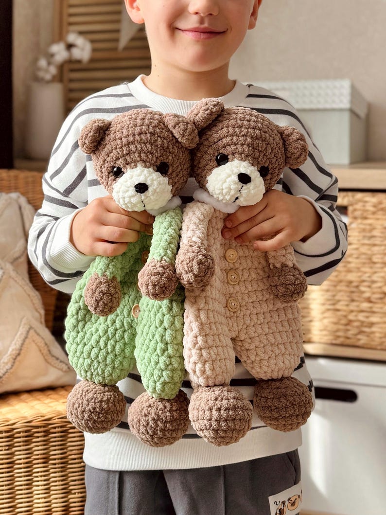 May include: Two crocheted teddy bears wearing matching outfits. The bear on the left is wearing a green outfit and the bear on the right is wearing a beige outfit. Both bears have brown fur and white faces.