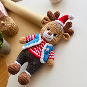 May include: A handmade crochet reindeer doll wearing a red and white sweater, blue vest, and a Santa hat. The reindeer has brown antlers, legs, and feet. The doll is a festive holiday decoration.