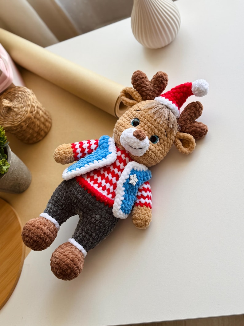 May include: A handmade crochet reindeer doll wearing a red and white sweater, blue vest, and a Santa hat. The reindeer has brown antlers, legs, and feet. The doll is a festive holiday decoration.