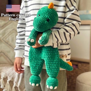 May include: A green crochet dinosaur toy with white accents on its feet and a small orange horn. The dinosaur is held by a person wearing a striped shirt. The text "Pattern crochet English" is visible.