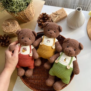 May include: Three handmade crochet teddy bears in a woven basket. Each bear wears a different colored pair of overalls: red, yellow, and green. The overalls have wooden buttons and are paired with white shirts. The bears are brown.