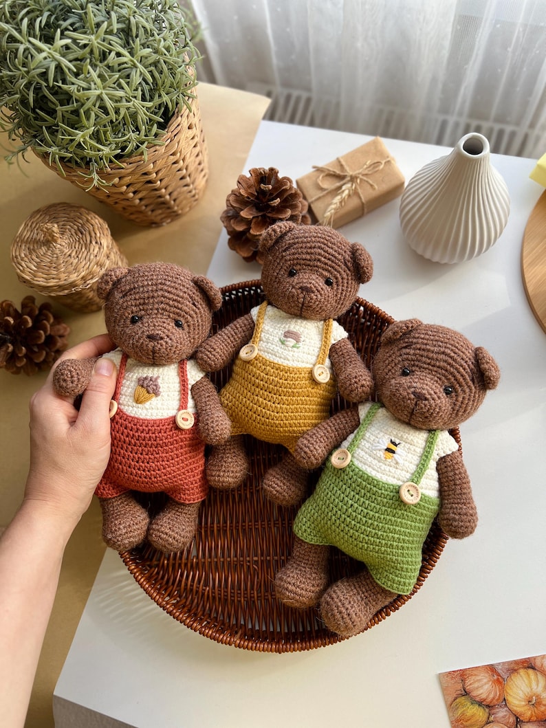 May include: Three handmade crochet teddy bears in a woven basket. Each bear wears a different colored pair of overalls: red, yellow, and green. The overalls have wooden buttons and are paired with white shirts. The bears are brown.