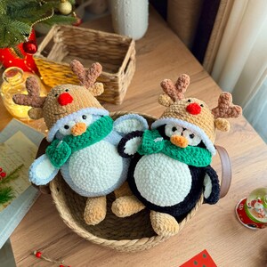 May include: Two handmade crochet penguin ornaments, each wearing a reindeer antler hat and a green scarf, sit in a woven basket. One penguin is blue and white, the other is black and white. A Christmas tree and holiday cards are in the background.