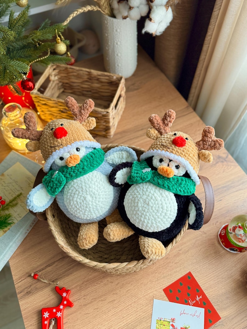 May include: Two handmade crochet penguin ornaments, each wearing a reindeer antler hat and a green scarf, sit in a woven basket. One penguin is blue and white, the other is black and white. A Christmas tree and holiday cards are in the background.