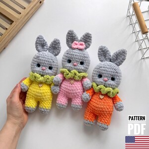 May include: Three crocheted gray bunny plushies wearing different colored outfits. The bunny on the left is wearing a yellow outfit, the middle bunny is wearing a pink outfit, and the bunny on the right is wearing an orange outfit. Each bunny has a green crocheted collar with a flower accent.