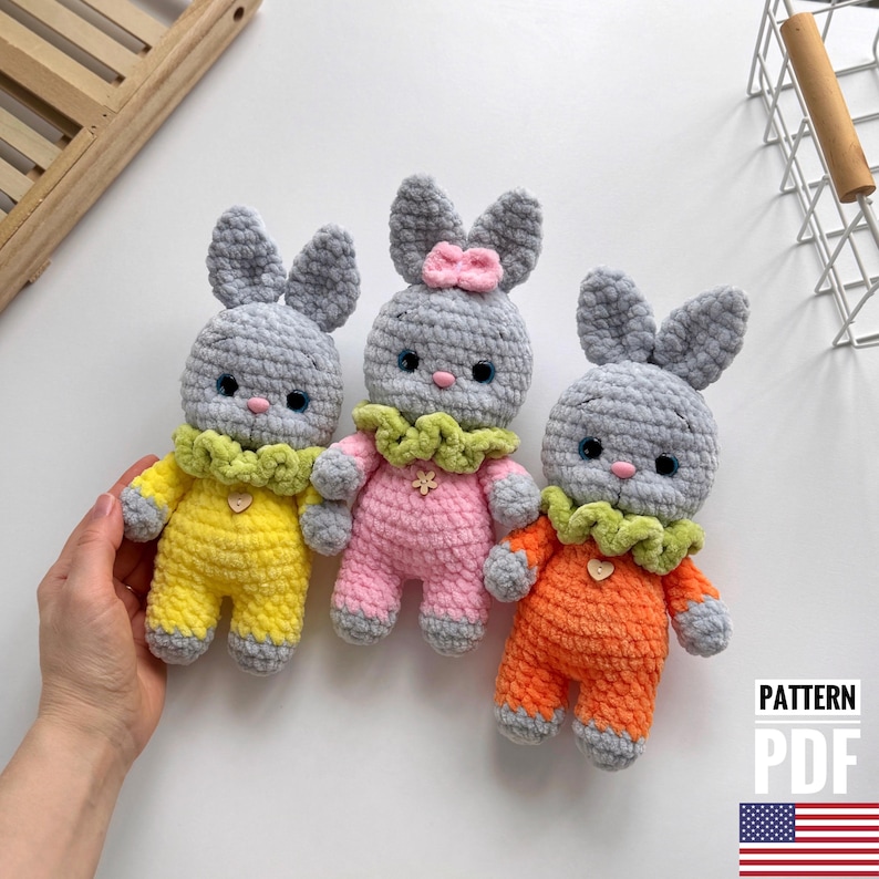 May include: Three crocheted gray bunny plushies wearing different colored outfits. The bunny on the left is wearing a yellow outfit, the middle bunny is wearing a pink outfit, and the bunny on the right is wearing an orange outfit. Each bunny has a green crocheted collar with a flower accent.