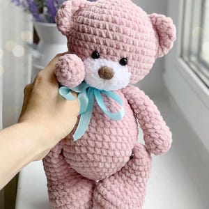 May include: A pink crocheted teddy bear with a white face and blue ribbon. The bear has black eyes and a brown nose.